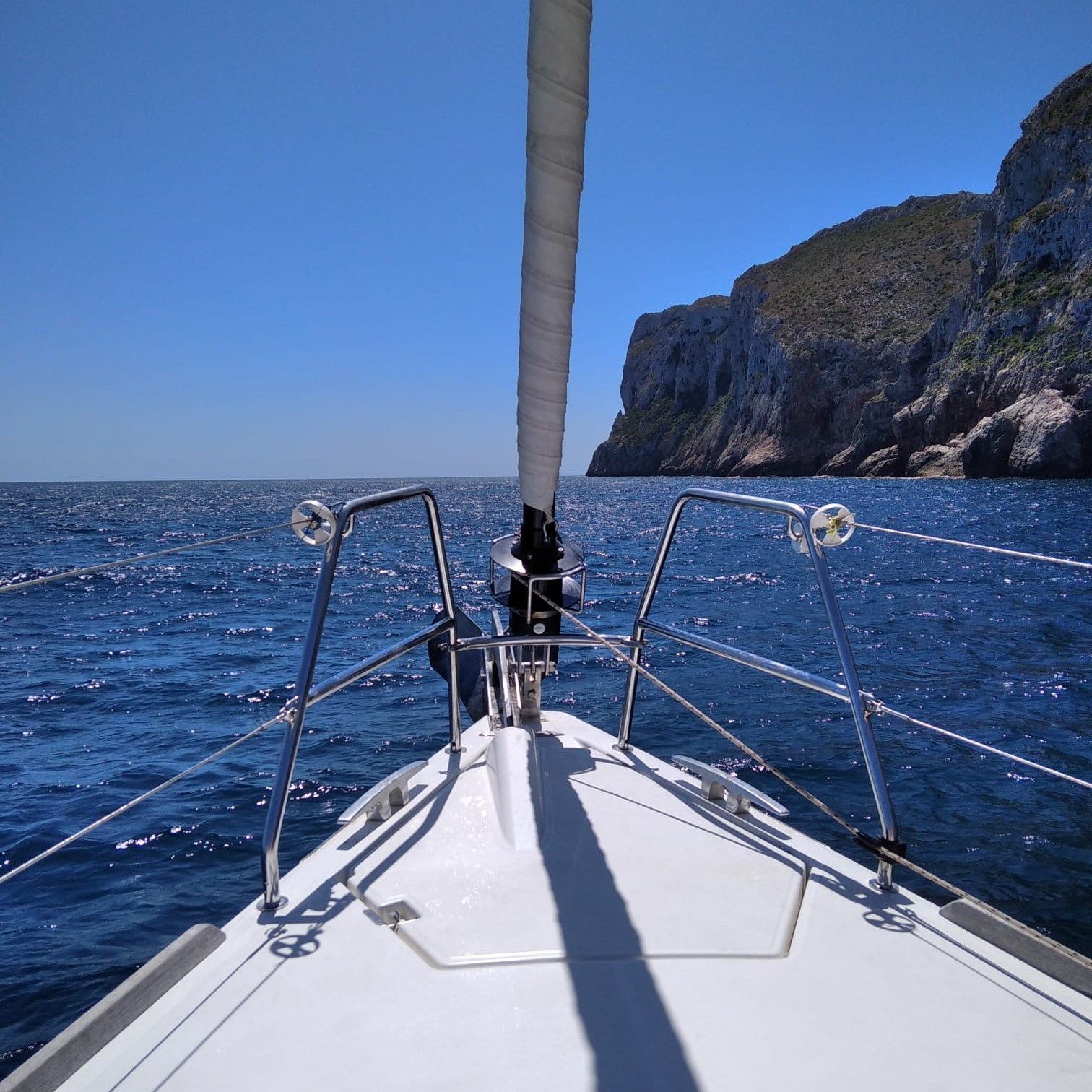 Vista desde la proa de un velero en el mar azul, acercándose a una costa rocosa bajo un cielo despejado.