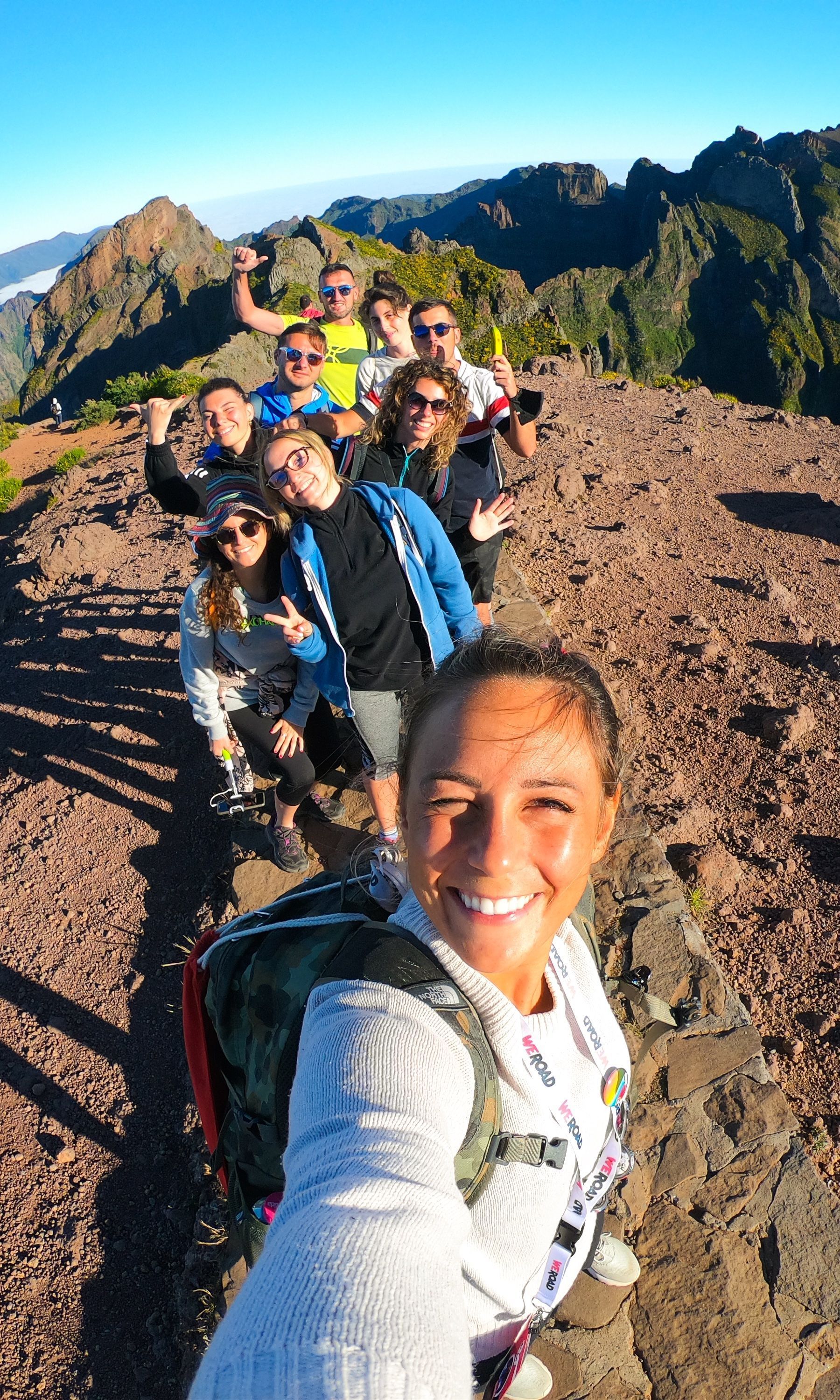A smiling WeRoad group trip poses for a selfie while hiking along a sunny, narrow mountain ridge.