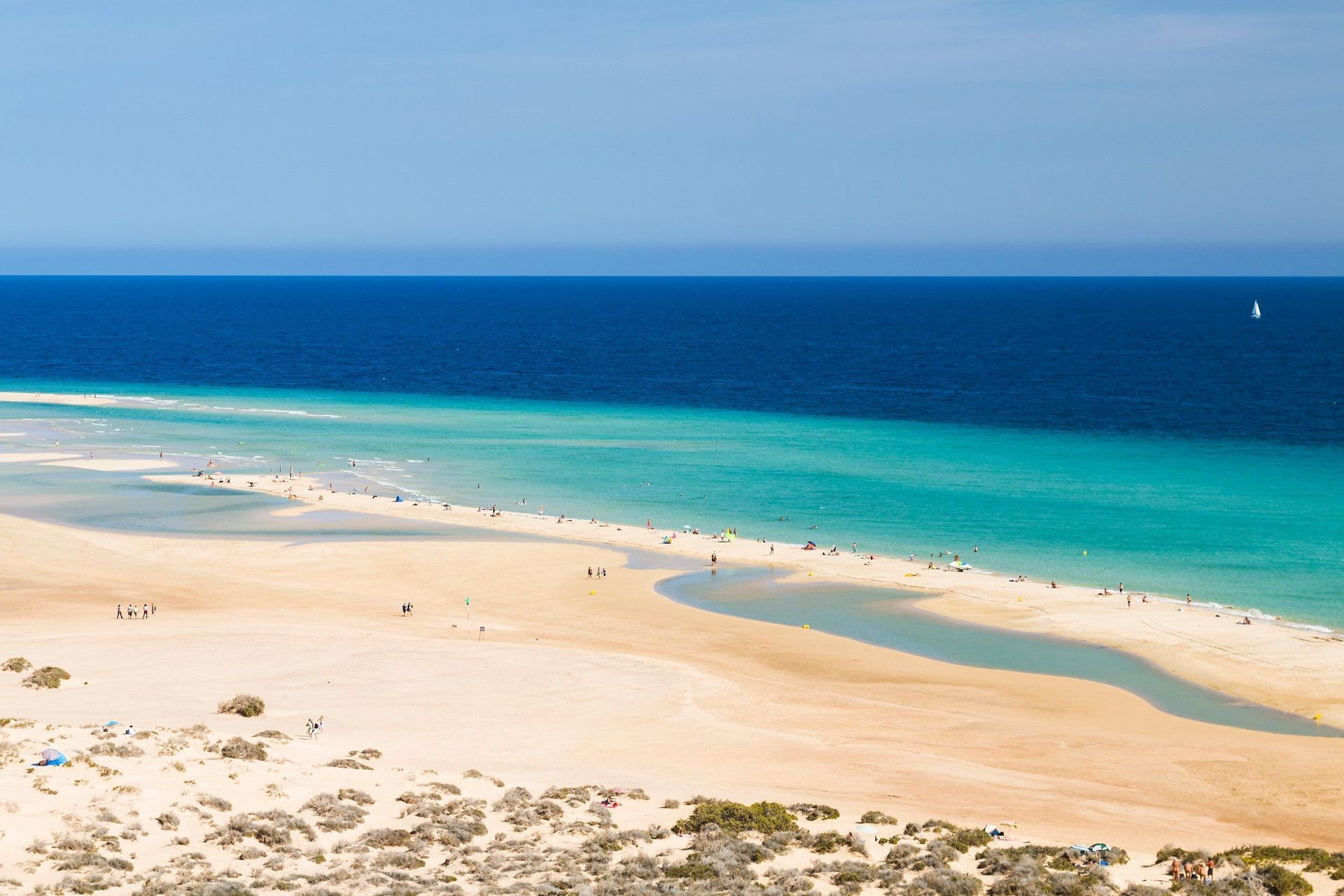 Una vasta spiaggia sabbiosa con persone lungo la riva dove l'acqua turchese incontra l'oceano blu profondo.