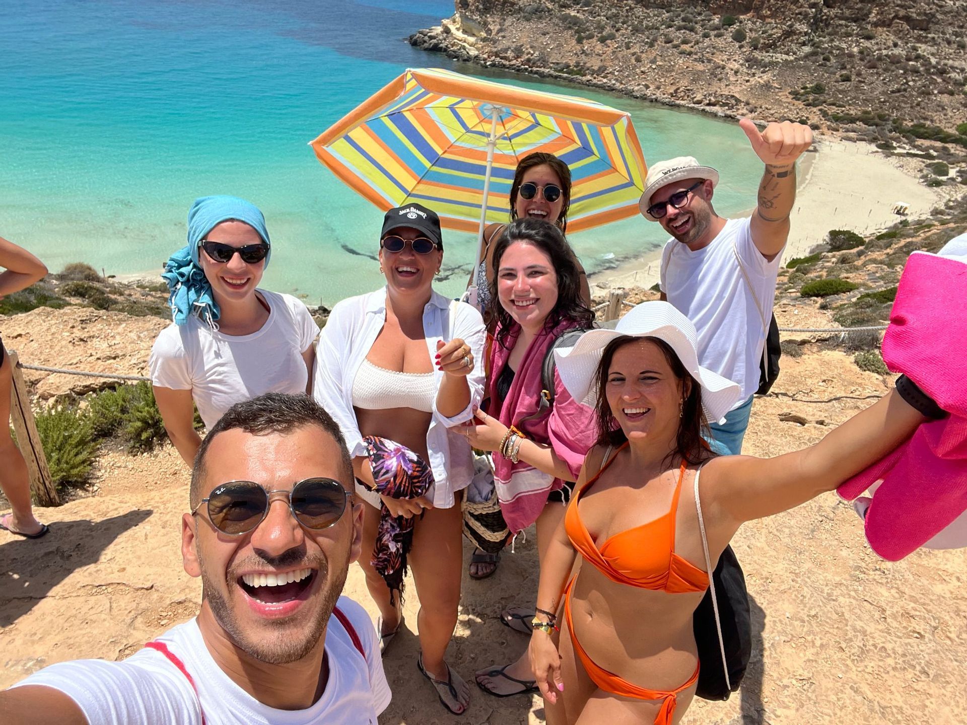 Un gruppo WeRoad scatta un selfie sorridente su una scogliera a picco su una spiaggia con acqua turchese.