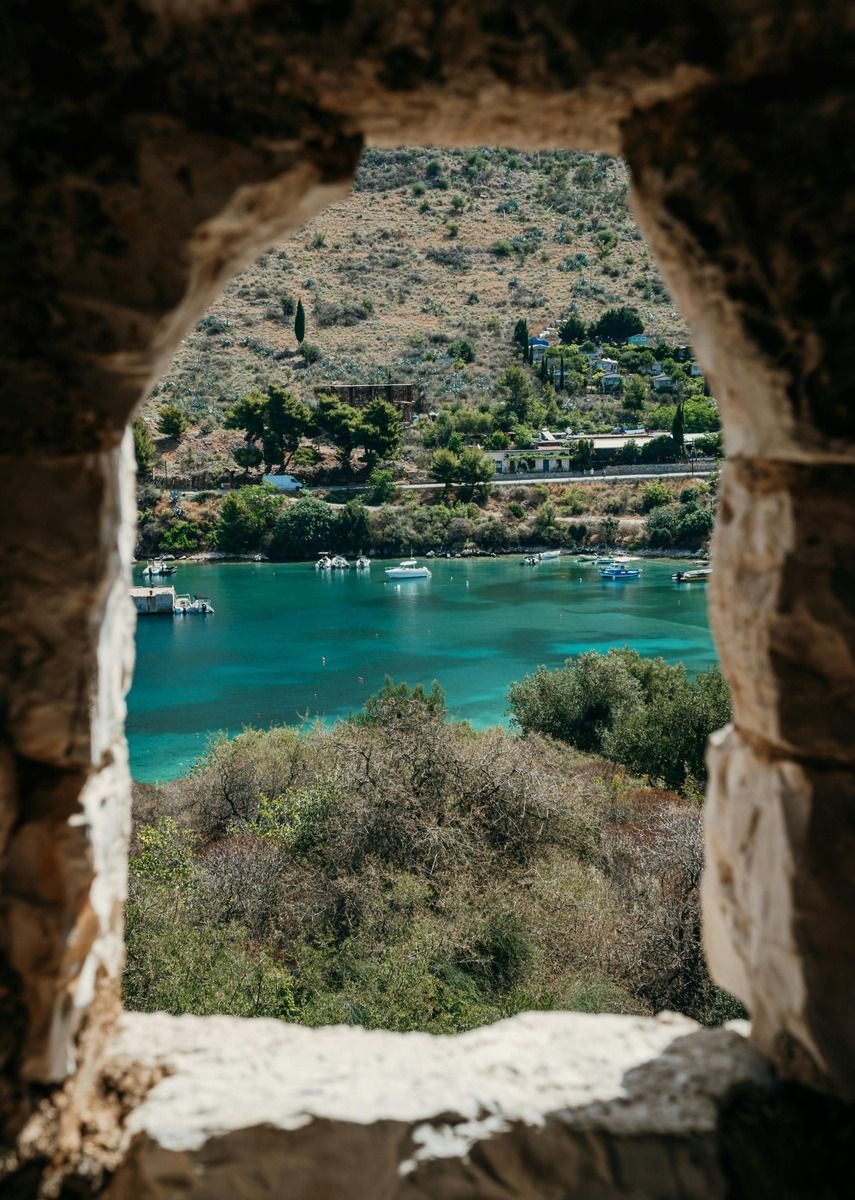 Una vista attraverso una finestra di pietra che si affaccia su una baia con acqua turchese, piccole imbarcazioni e una costa verde e collinare.