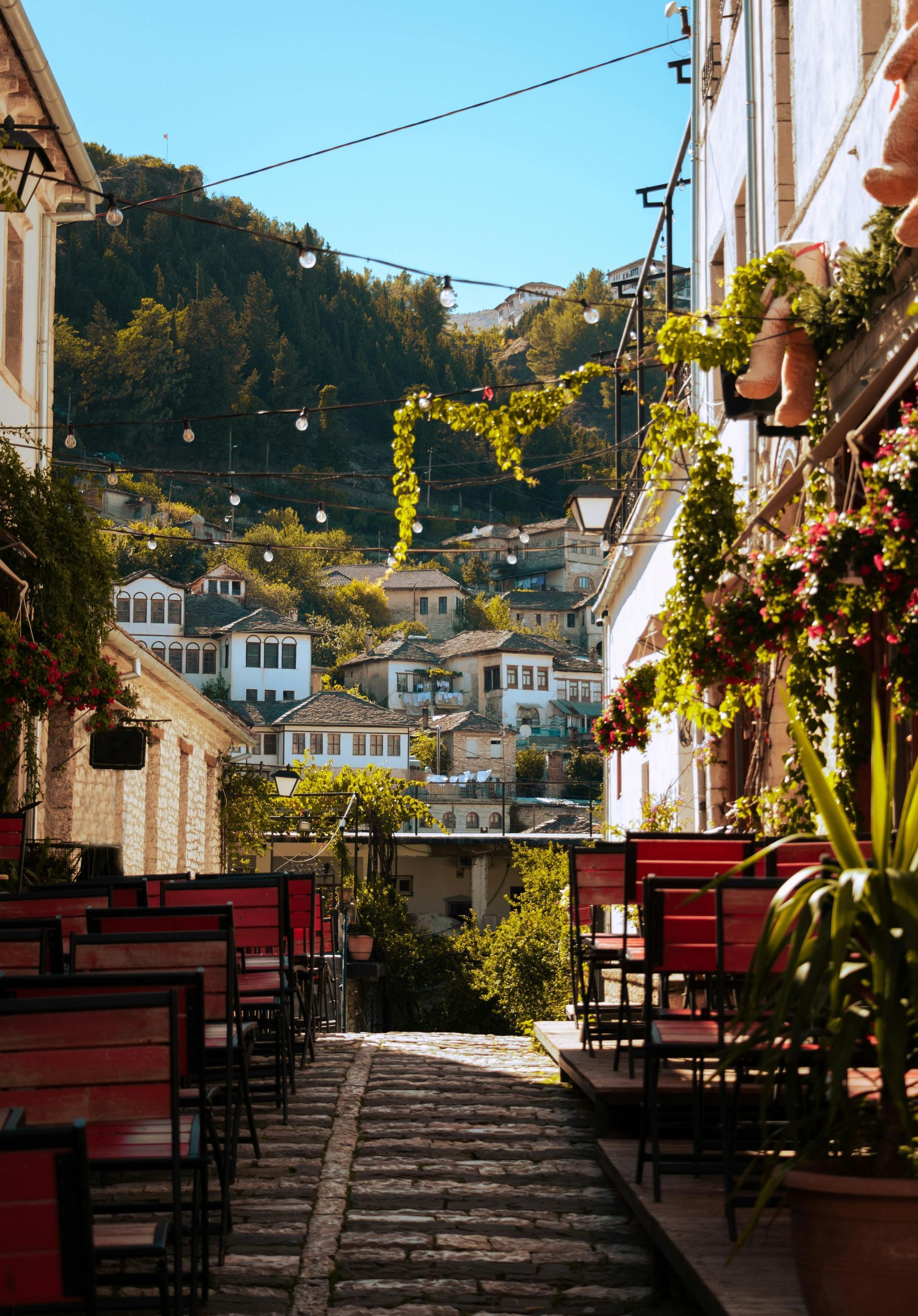 A cobblestone alley lined with red chairs from an outdoor cafe ascends a hill with stone houses and string lights.