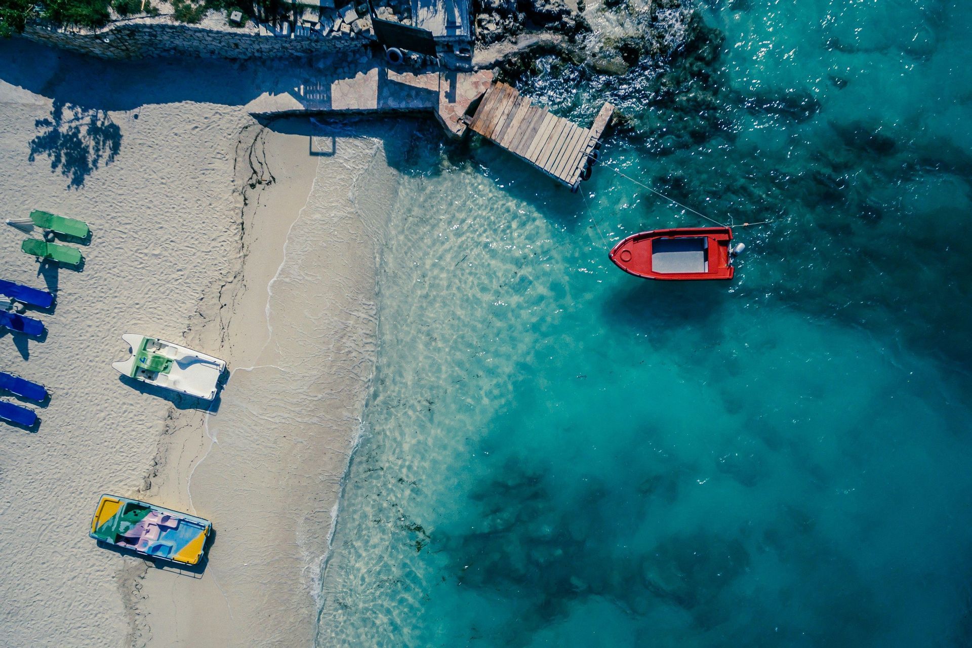 Vista aerea dall'alto di una spiaggia sabbiosa con diverse barche sulla sabbia e una barca rossa che galleggia in acqua turchese vicino a un molo.