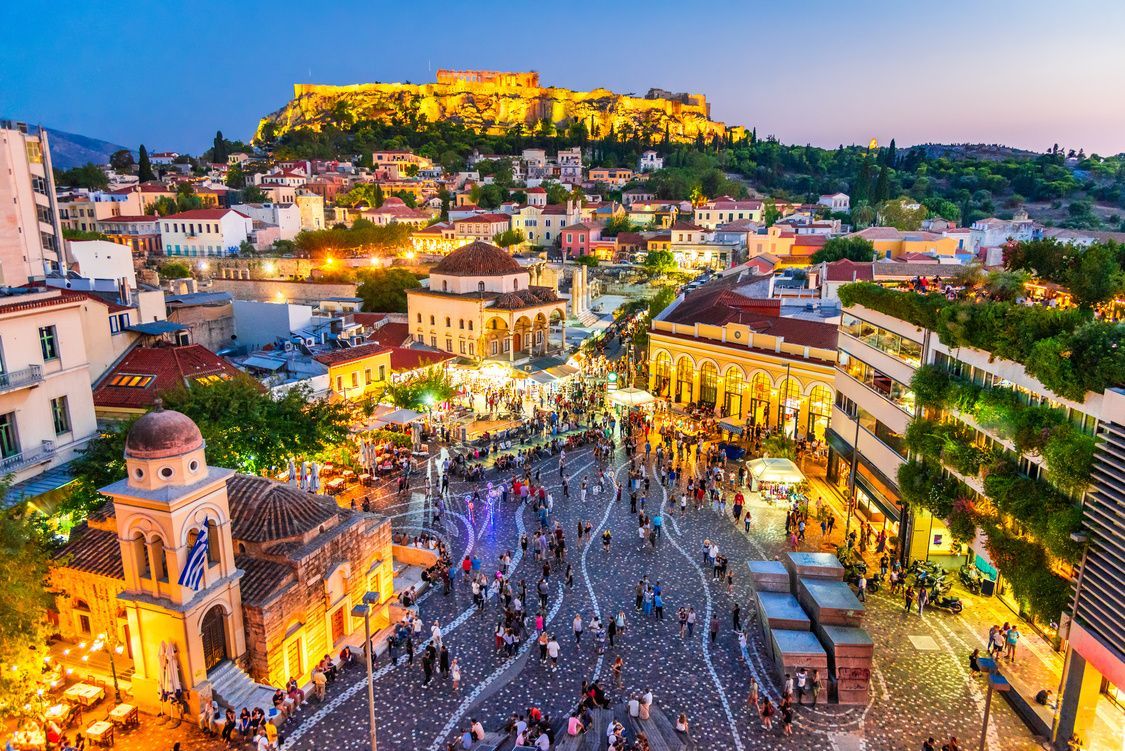 Una veduta sopraelevata di una piazza cittadina affollata al crepuscolo, con edifici storici illuminati e un'acropoli illuminata su una collina sullo sfondo.