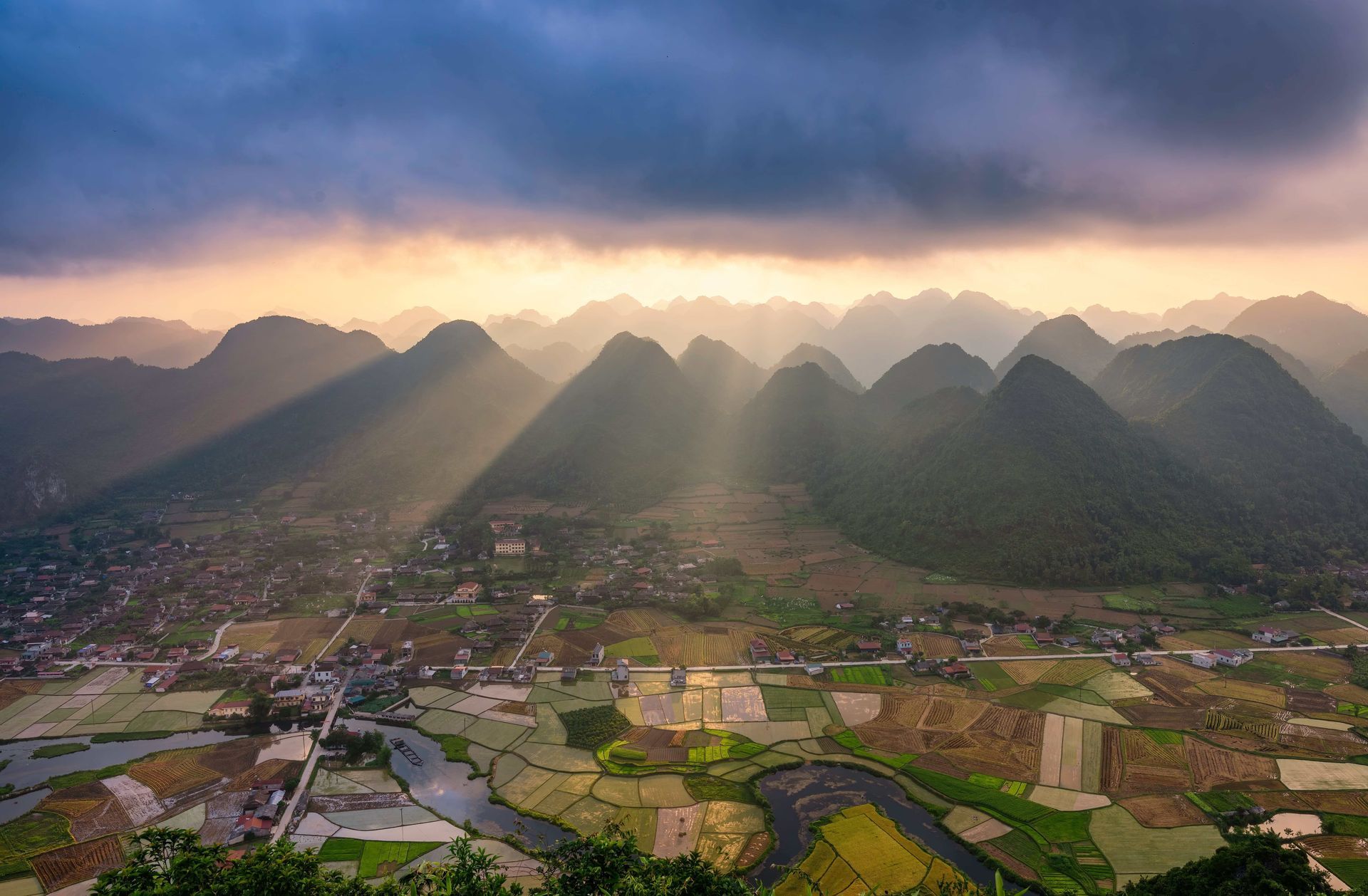 Des rayons de soleil percent les nuages sombres sur une vallée de montagnes vertes, des rizières en patchwork et un petit village.