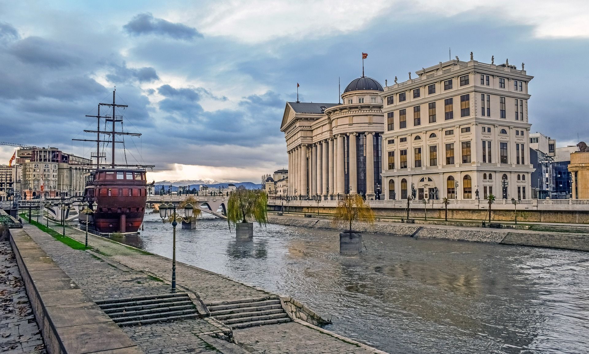 Una vista lungo il fiume di una città con una nave di legno attraccata accanto a un grande edificio neoclassico sotto un cielo nuvoloso.