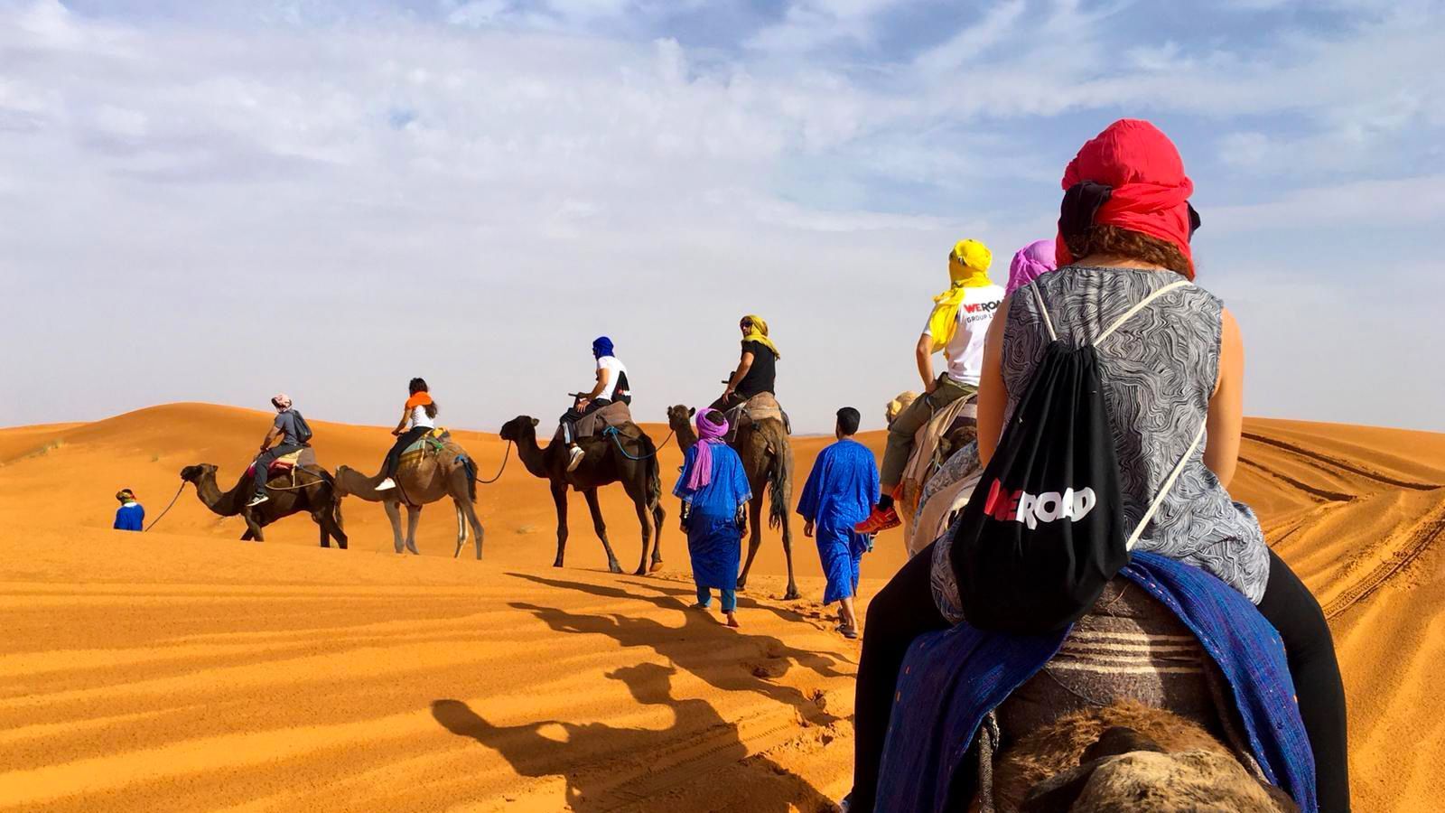 Un viaggio di gruppo WeRoad cavalca cammelli in fila attraverso vaste dune di sabbia arancioni sotto un cielo parzialmente nuvoloso.