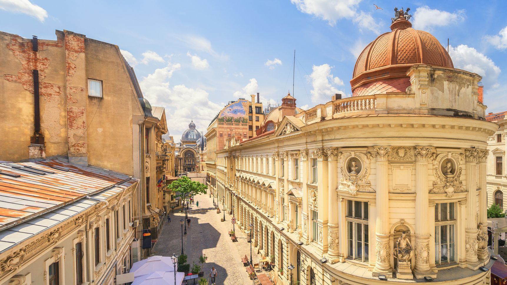 Vista en picada de una calle de ciudad soleada, bordeada por edificios clásicos ornamentados y una gran cúpula naranja bajo un cielo azul.