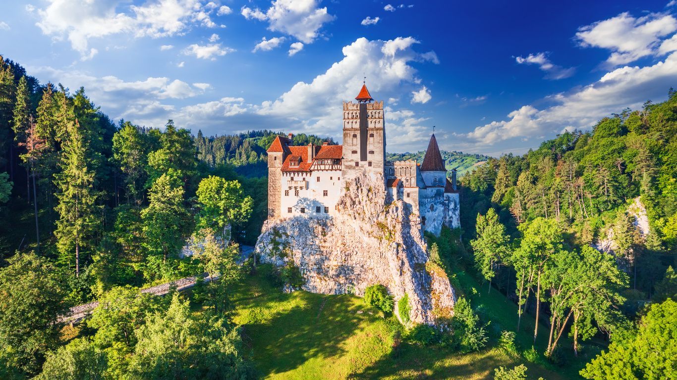 Ein weißes Schloss mit roten Ziegeldächern thront auf einer felsigen Klippe, umgeben von einem dichten grünen Wald unter einem blauen Himmel mit Wolken.