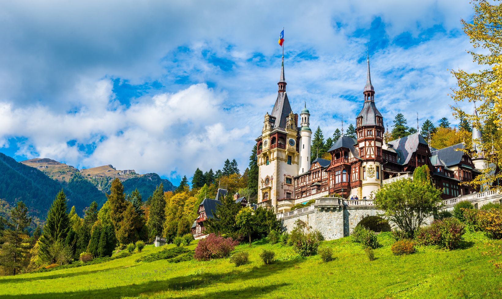 Un castillo grande y ornamentado con múltiples torres se asienta en una colina verde y cubierta de hierba, respaldado por un denso bosque y montañas bajo un cielo azul y nublado.