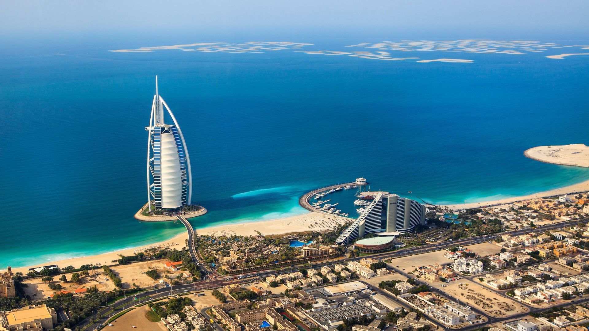 Vue aérienne d'un hôtel en forme de voile sur une petite île artificielle, relié par un pont à un littoral sablonneux et à une ville.
