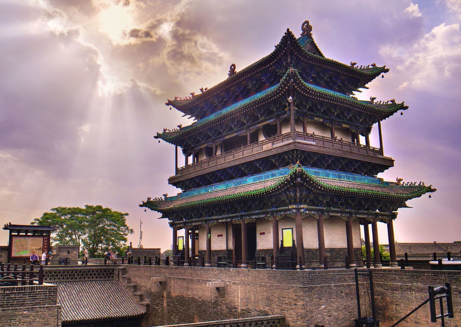 Eine traditionelle chinesische Pagode mit grünen Ziegeldächern thront auf einer Steinmauer unter einem Himmel mit Wolken und durchdringenden Sonnenstrahlen.