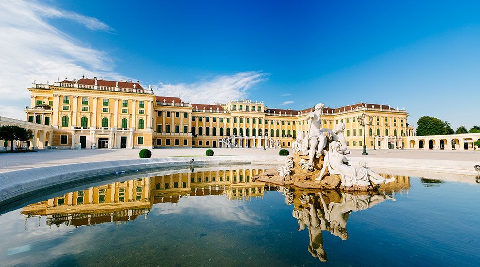 A large yellow palace with green shutters stands behind a fountain with white marble statues, reflected in the water on a clear day.
