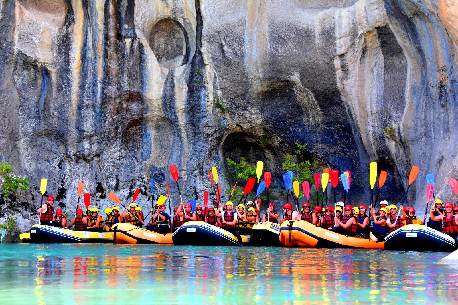 Un viaggio di gruppo WeRoad in rafting solleva pagaie colorate su un fiume turchese davanti a un'imponente parete rocciosa.