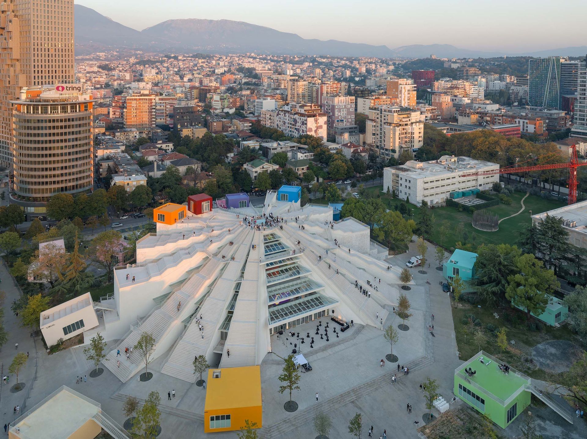 Une vue aérienne d'un bâtiment moderne, blanc, en forme de pyramide, avec des boîtes colorées sur son toit, situé dans une ville animée au coucher du soleil.
