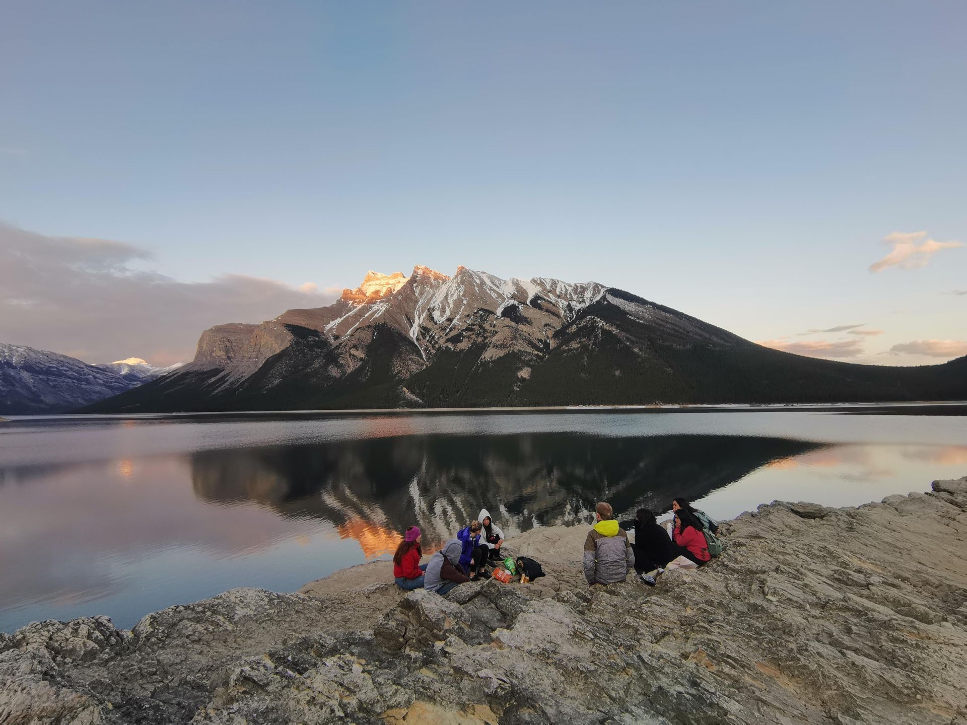 Un gruppo WeRoad su una riva rocciosa, ammira il tramonto che illumina una montagna innevata riflessa in un lago calmo.