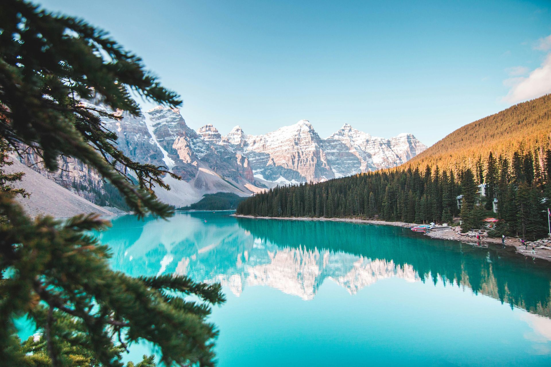 Montagnes enneigées et forêt de pins dense se reflétant dans l'eau calme et turquoise d'un lac sous un ciel clair.