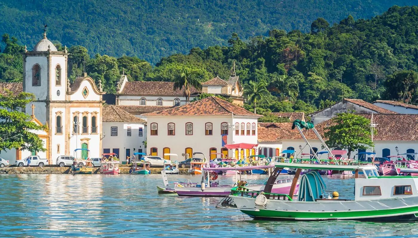 Colorful boats float in a bay in front of a coastal town with colonial-style buildings and a church, with a lush, green hill in the background.