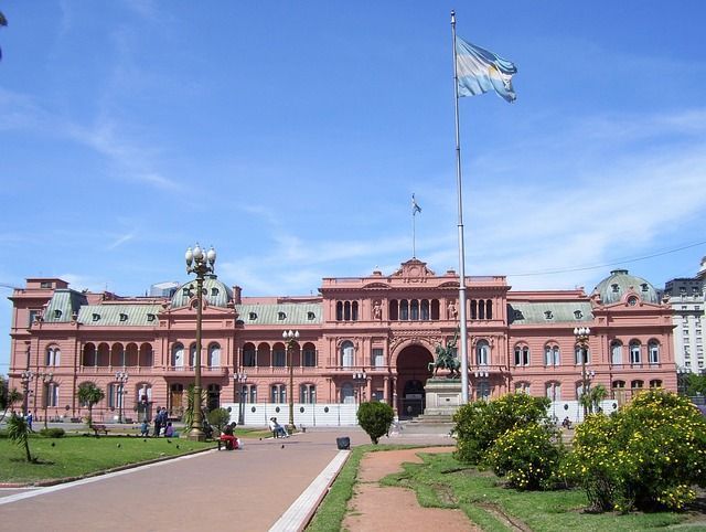 Un grande edificio governativo rosa si staglia dietro un parco, con una bandiera argentina che sventola su un'alta asta sotto un cielo azzurro intenso.