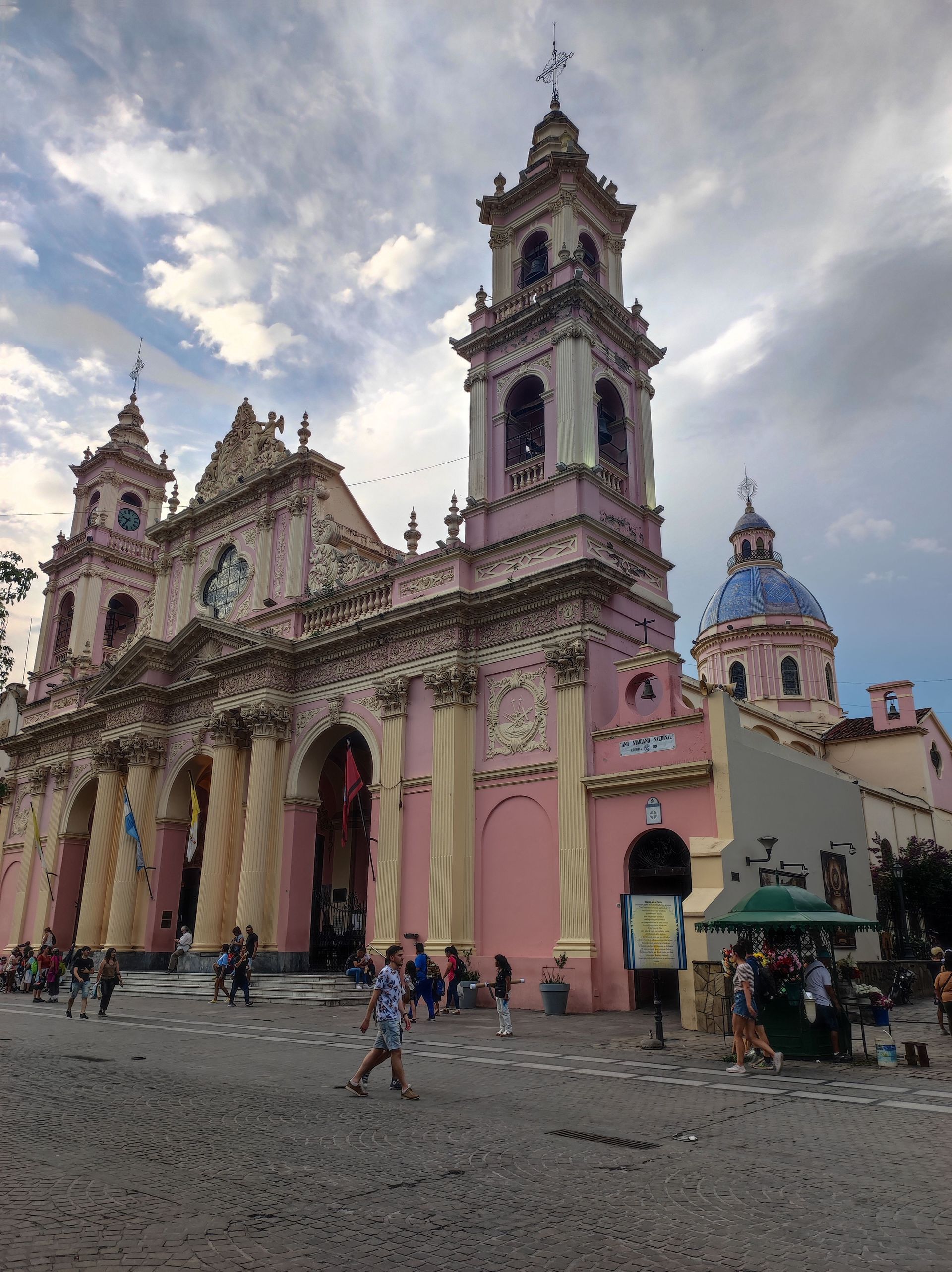 Una cattedrale rosa riccamente decorata, con colonne color crema e due campanili gemelli, si erge su una piazza acciottolata mentre le persone passeggiano.