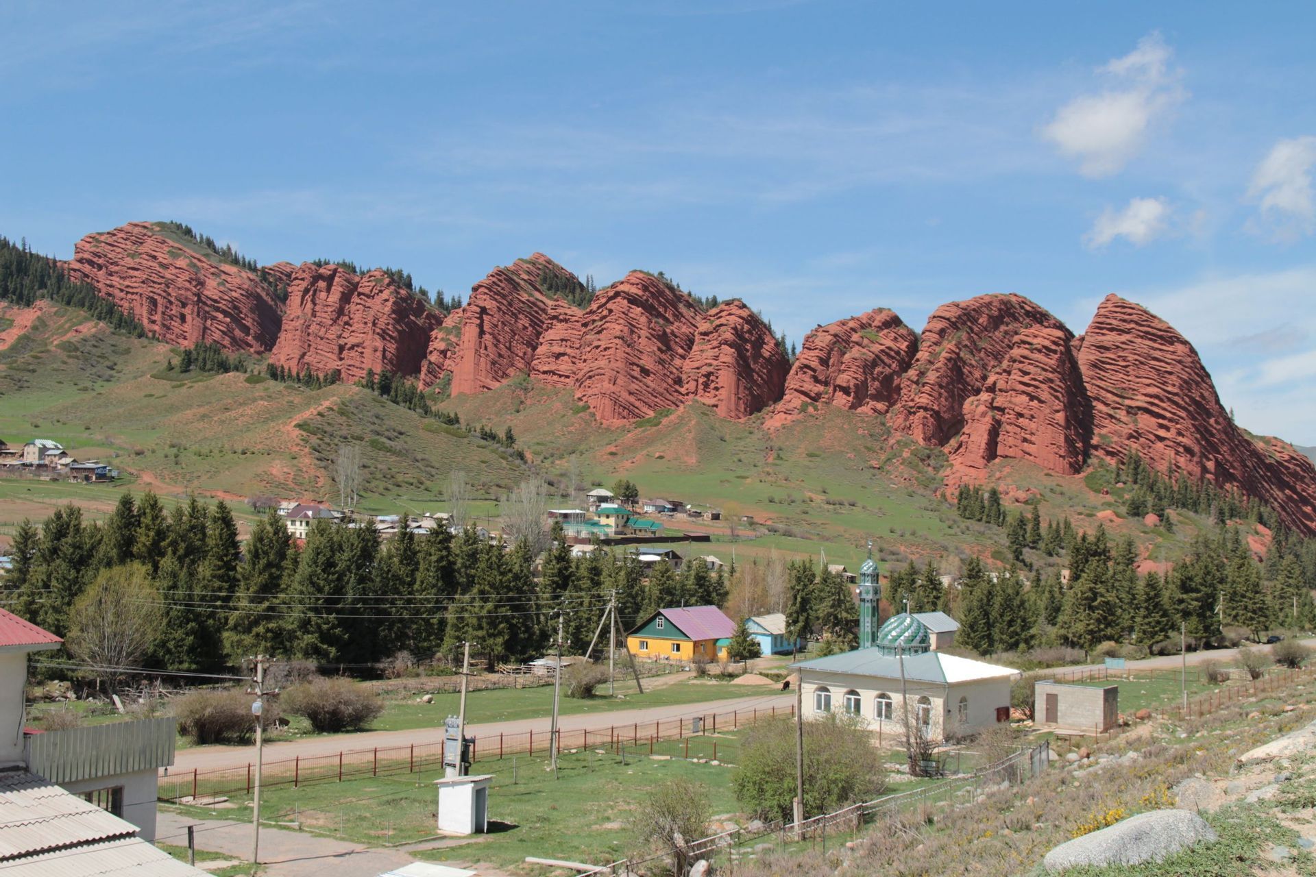 Un petit village avec une mosquée est niché au pied de grandes formations rocheuses rouges superposées, sur une colline verdoyante sous un ciel bleu.