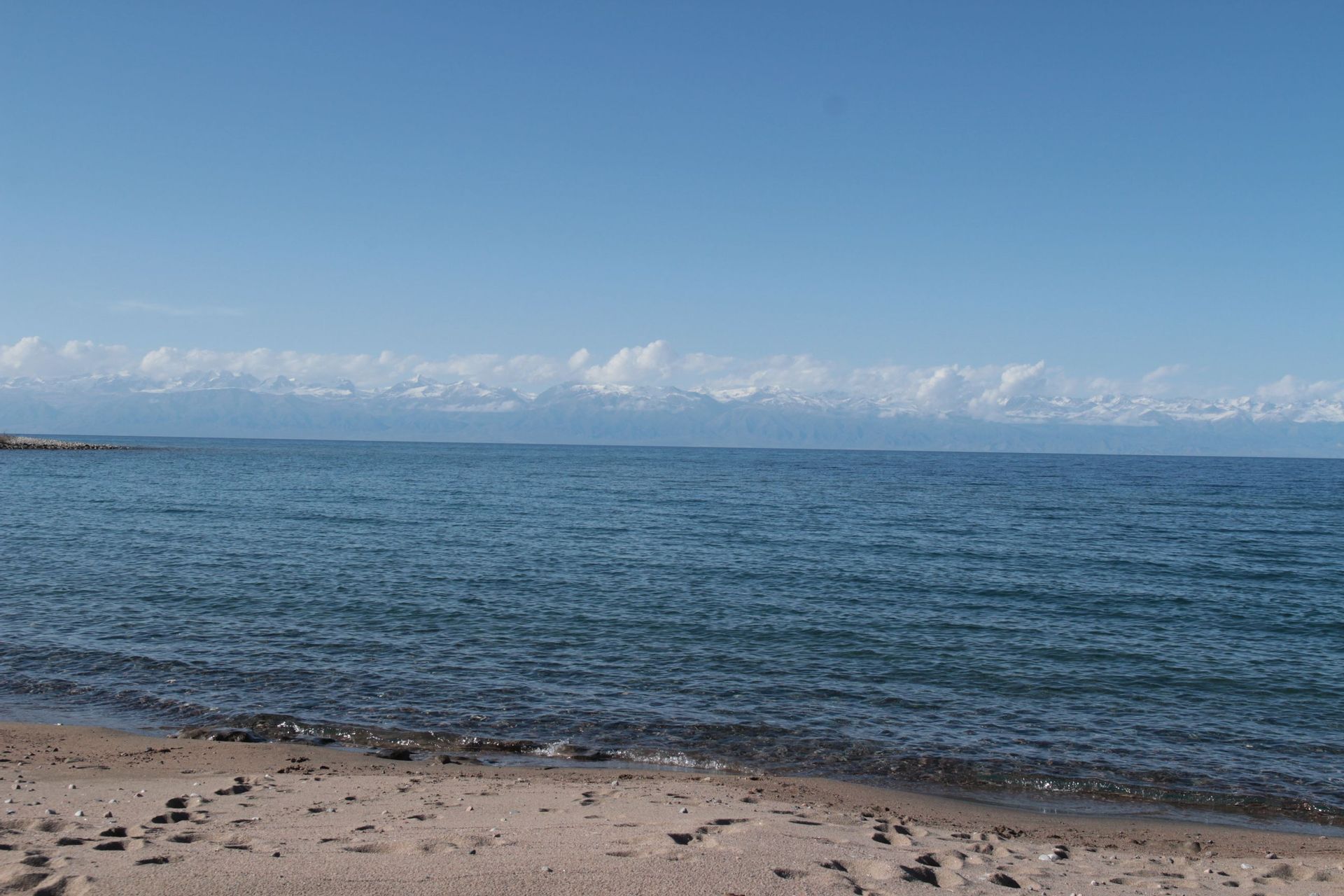 Une plage de sable rencontre l'eau bleue calme d'un lac, avec des montagnes enneigées visibles au loin sous un ciel clair.