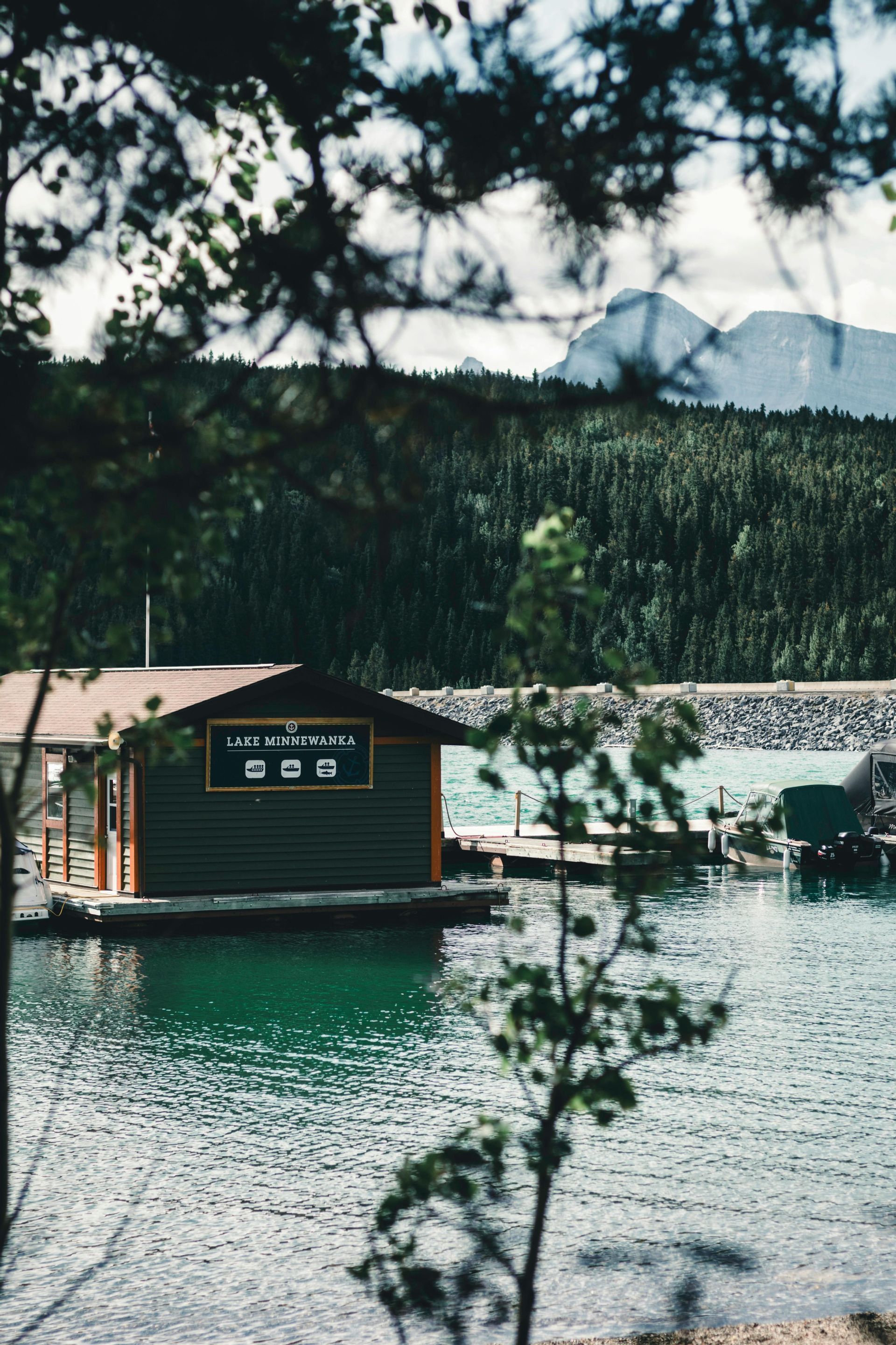 Un hangar à bateaux sur le lac Minnewanka avec un quai et des bateaux, encadré par des arbres avec une montagne et une forêt en arrière-plan.