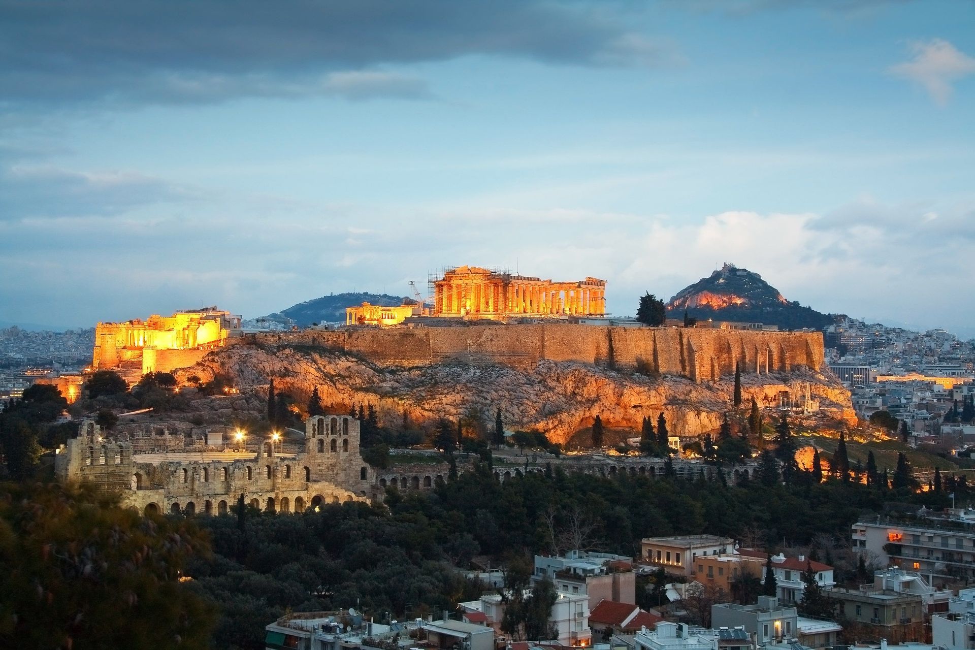 Un'antica cittadella, splendidamente illuminata da luci dorate, si erge su una collina rocciosa che domina un paesaggio urbano al tramonto.