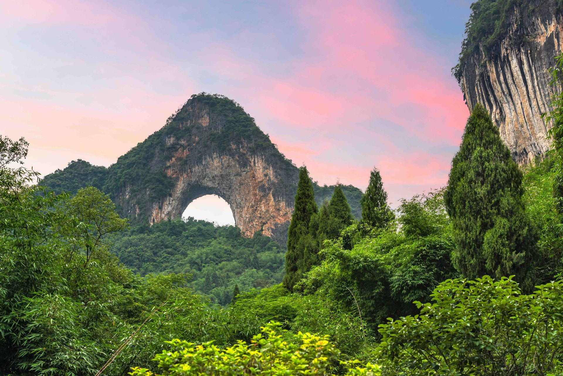 Une montagne avec une grande arche naturelle s'élève au-dessus d'une forêt verte dense sous un ciel rose et violet.