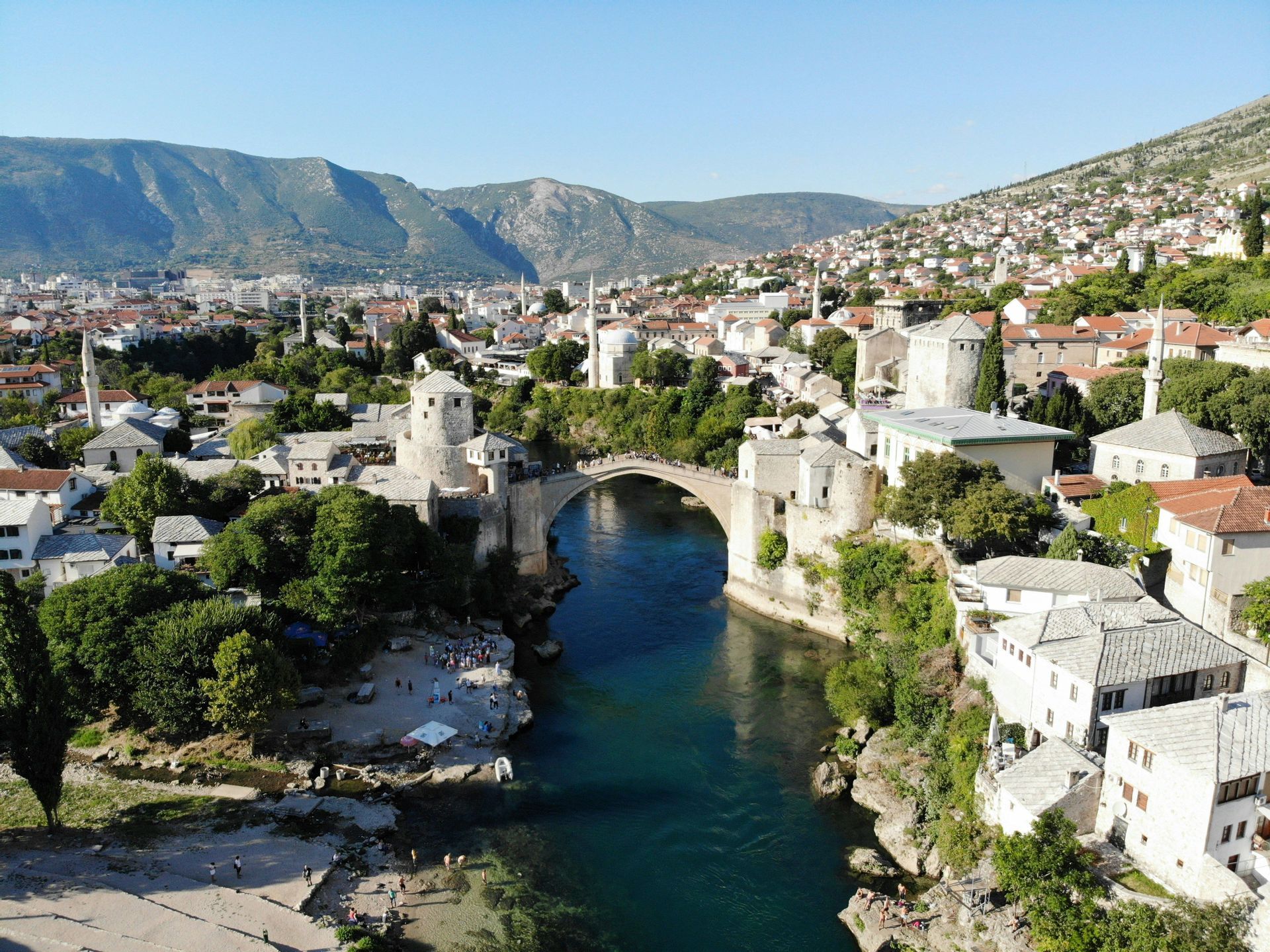 Una vista aerea di un vecchio ponte di pietra che attraversa un fiume turchese in una città storica, con case che si arrampicano sulle verdi colline sullo sfondo.