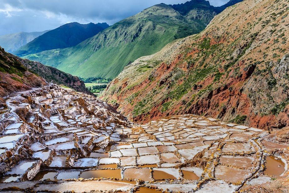 Terraced salt pans cascade down a mountainside, surrounded by lush green and reddish-brown slopes under a cloudy sky.
