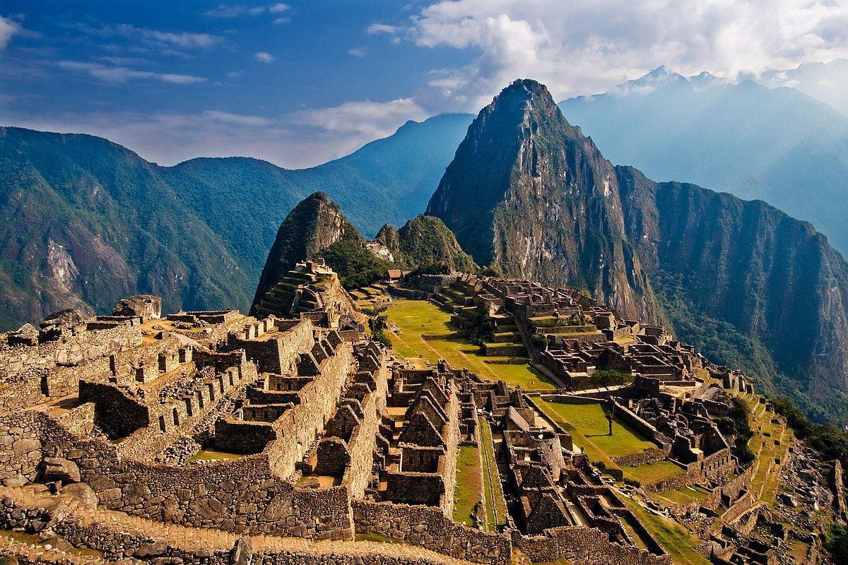 Ancient stone ruins on green terraces with tall, steep mountains in the background under a partly cloudy sky.