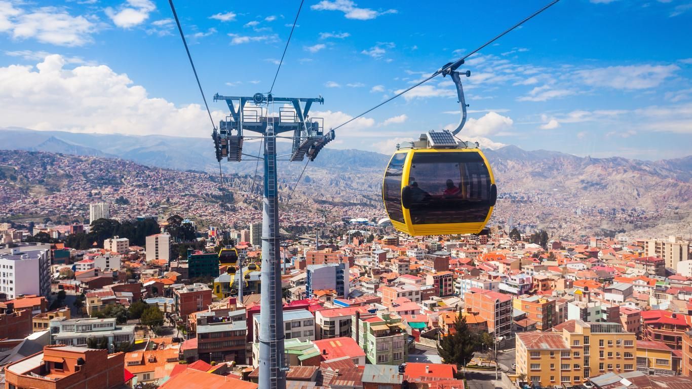 A yellow cable car travels on a line over a dense city at the foot of distant mountains under a cloudy blue sky.