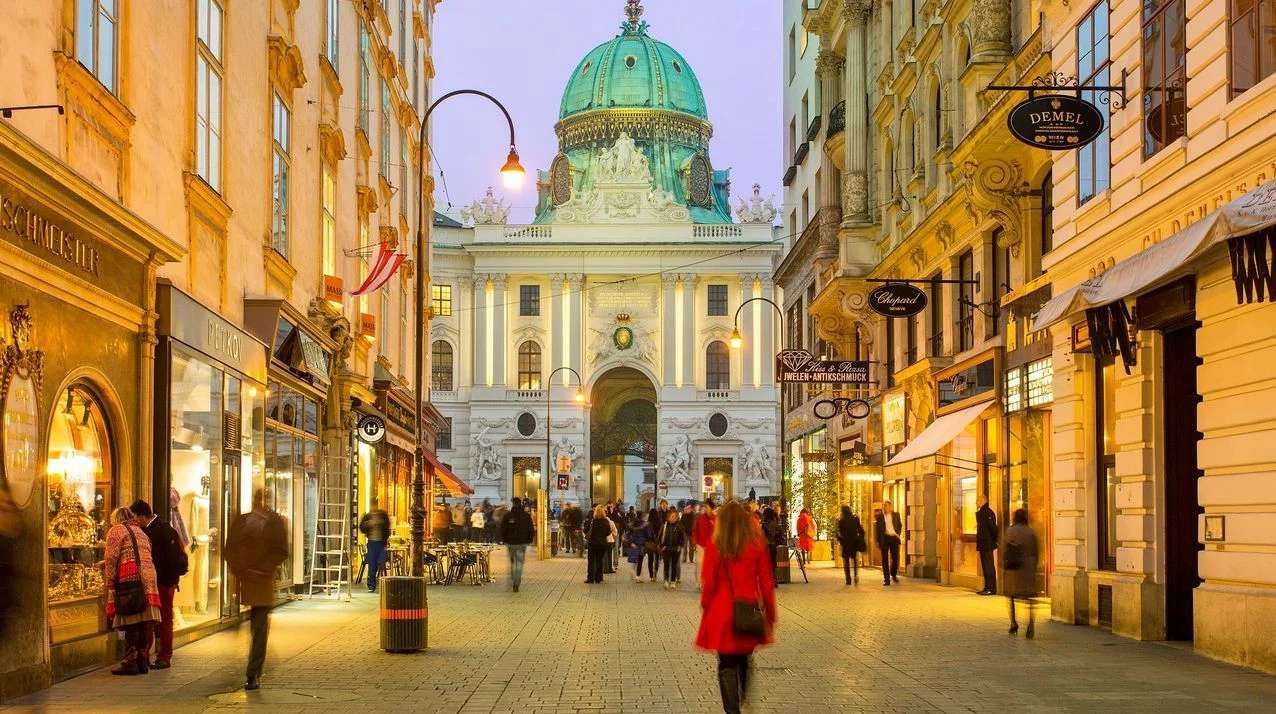 A busy cobblestone street at dusk, with people walking past illuminated shops towards a grand building with a green dome.