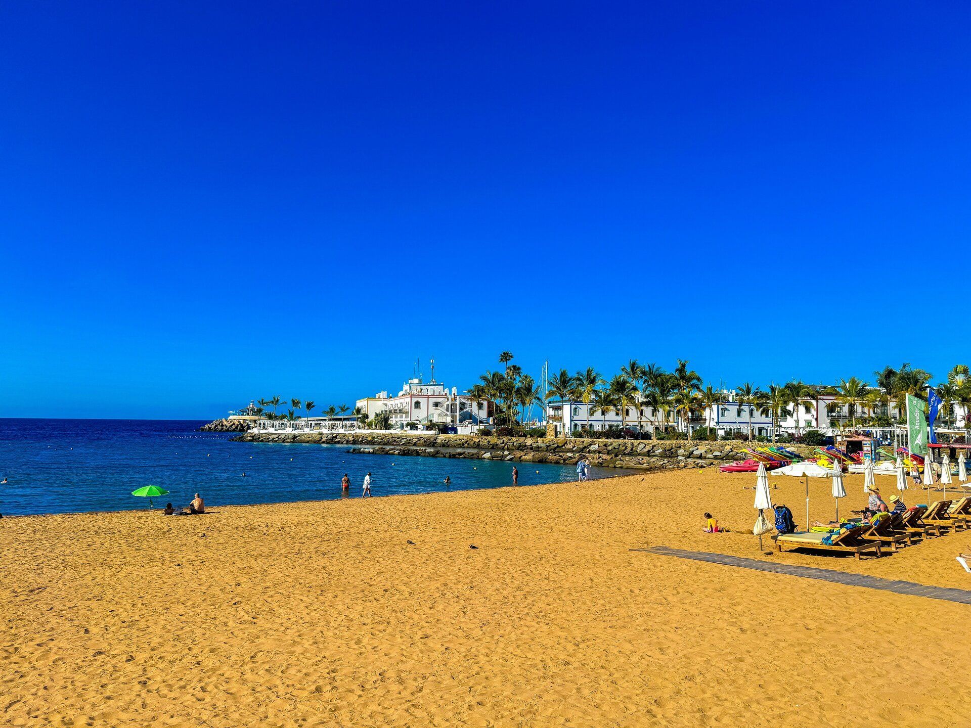 A wide, golden sand beach with people swimming and sunbathing by a calm blue sea, with white buildings and palm trees in the background.