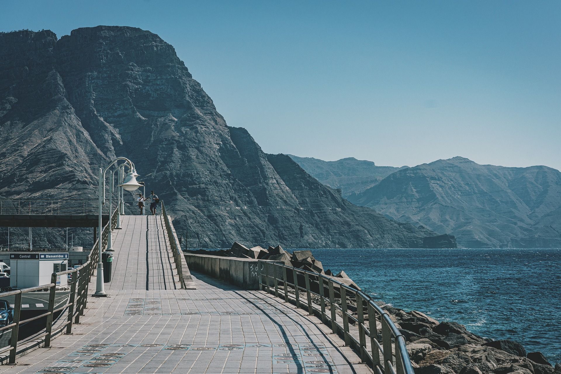 Two people on a WeRoad group trip walking along a paved seaside pier next to large, rocky mountains under a clear sky.
