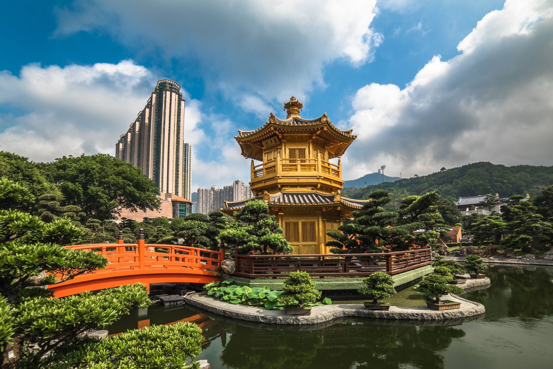 Une pagode dorée traditionnelle et un pont orange dans un bassin de jardin, avec une silhouette de ville moderne en arrière-plan sous un ciel bleu.