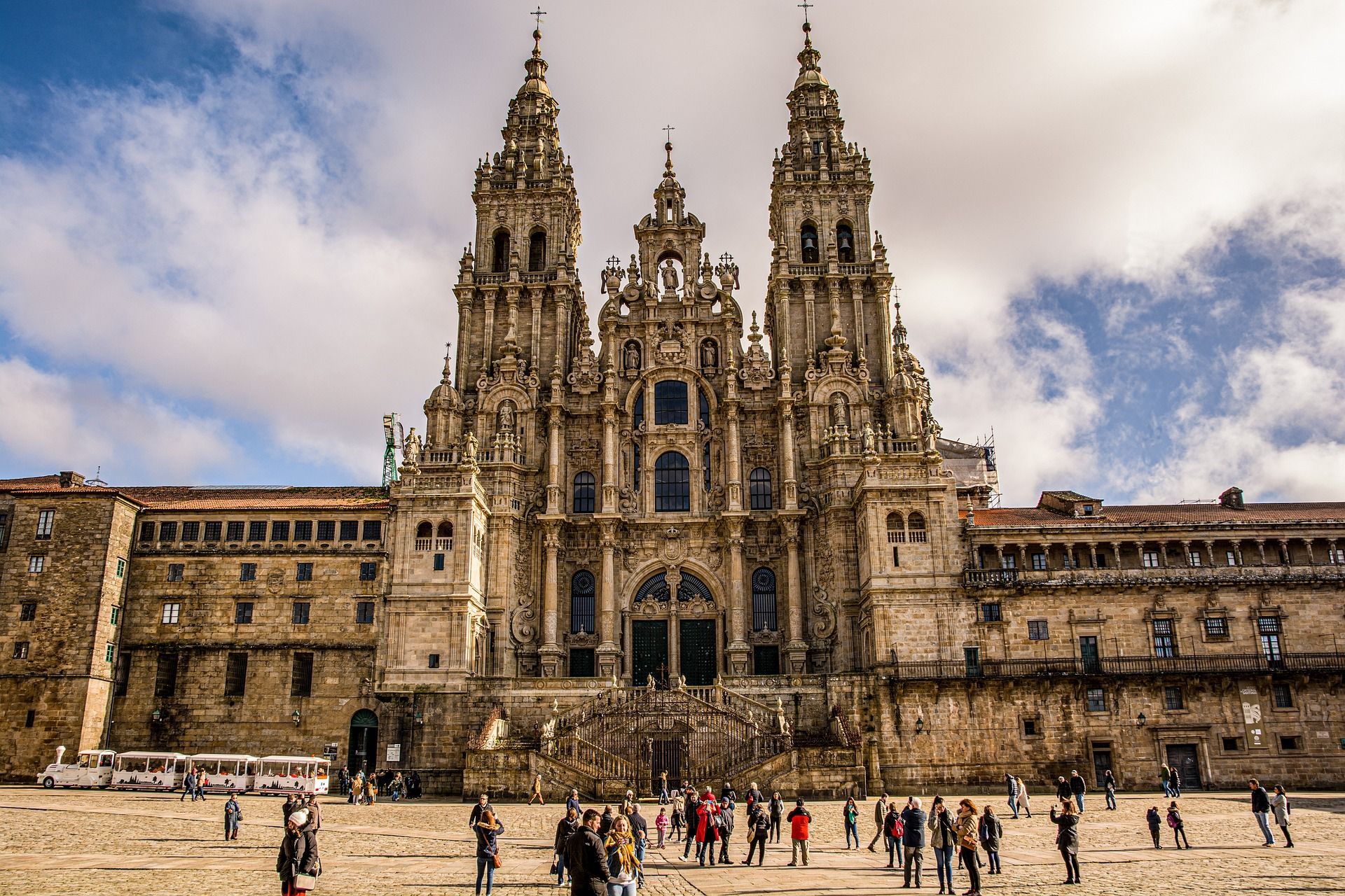 La ornamentada fachada de una gran catedral de piedra se eleva sobre una animada plaza llena de gente bajo un cielo parcialmente nublado.