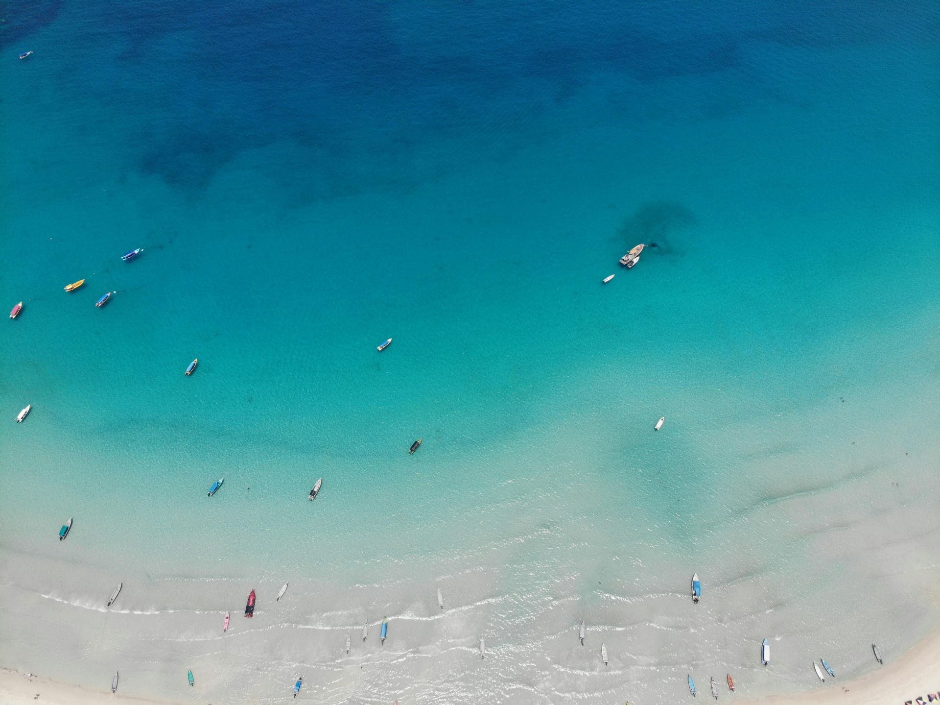 Luftaufnahme von vielen kleinen Booten, die auf einem weißen Sandstrand und im klaren türkisfarbenen Meer verstreut sind.