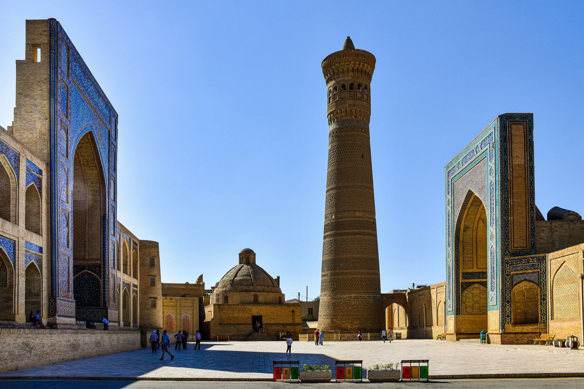 Un alto minareto in mattoni e sontuosi edifici con archi piastrellati di blu circondano una piazza in pietra soleggiata sotto un cielo limpido.