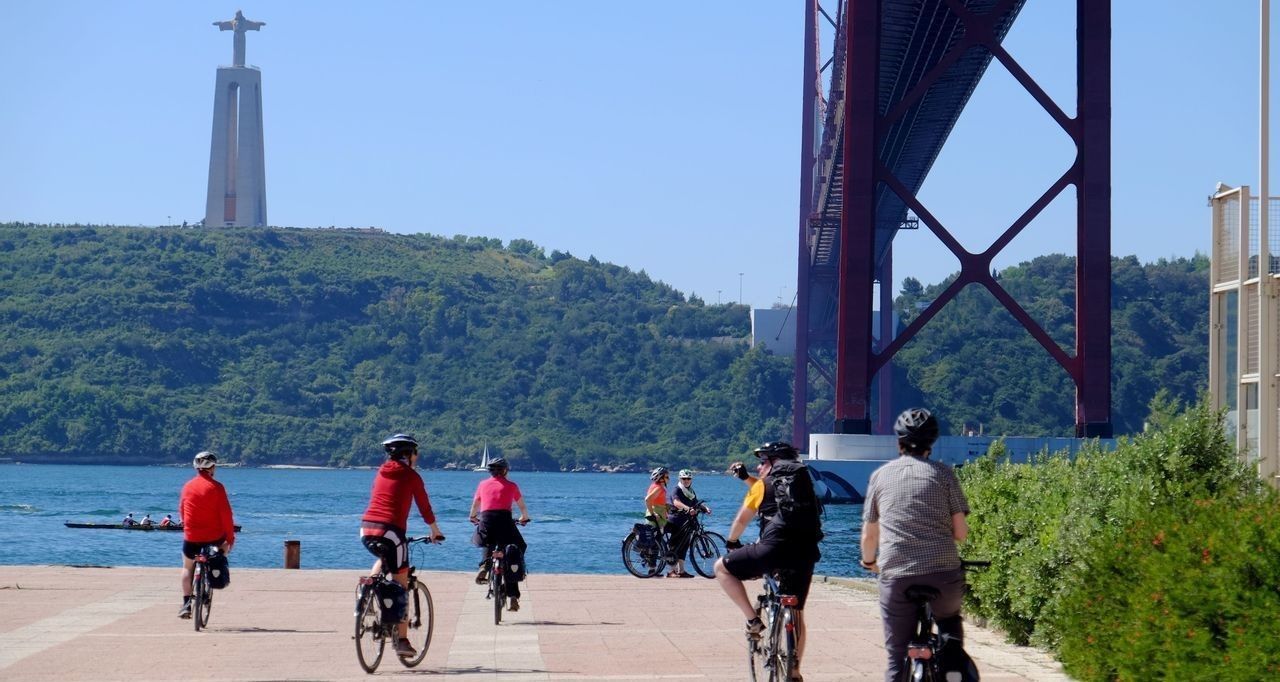 Un viaggio di gruppo WeRoad in bicicletta su un percorso lungomare con un grande ponte rosso e un monumento su una collina sullo sfondo.