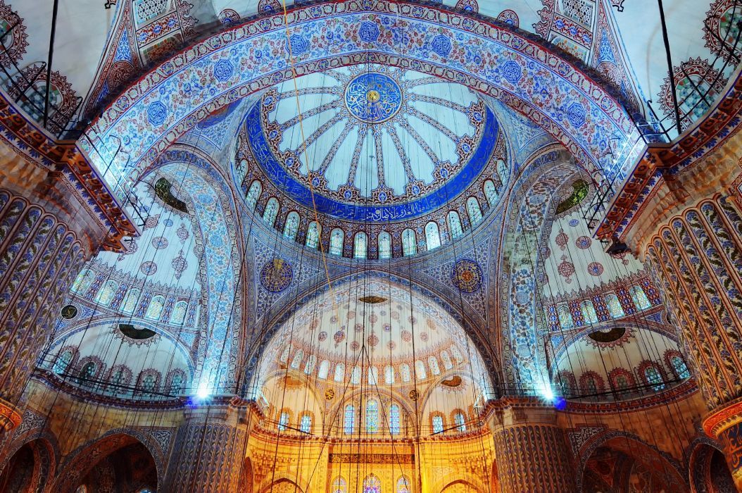 El interior de una mezquita con un techo abovedado intrincadamente decorado, con patrones azules y luces colgantes.