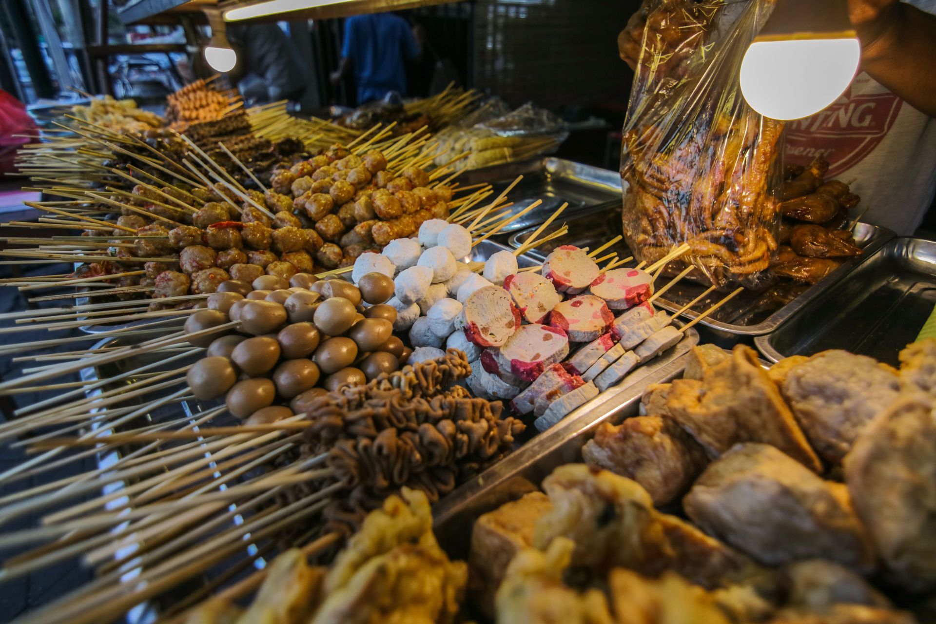 Variedad de brochetas de comida callejera, incluyendo huevos de codorniz, albóndigas y pasteles de pescado, exhibidas en bandejas de metal en un puesto de mercado.