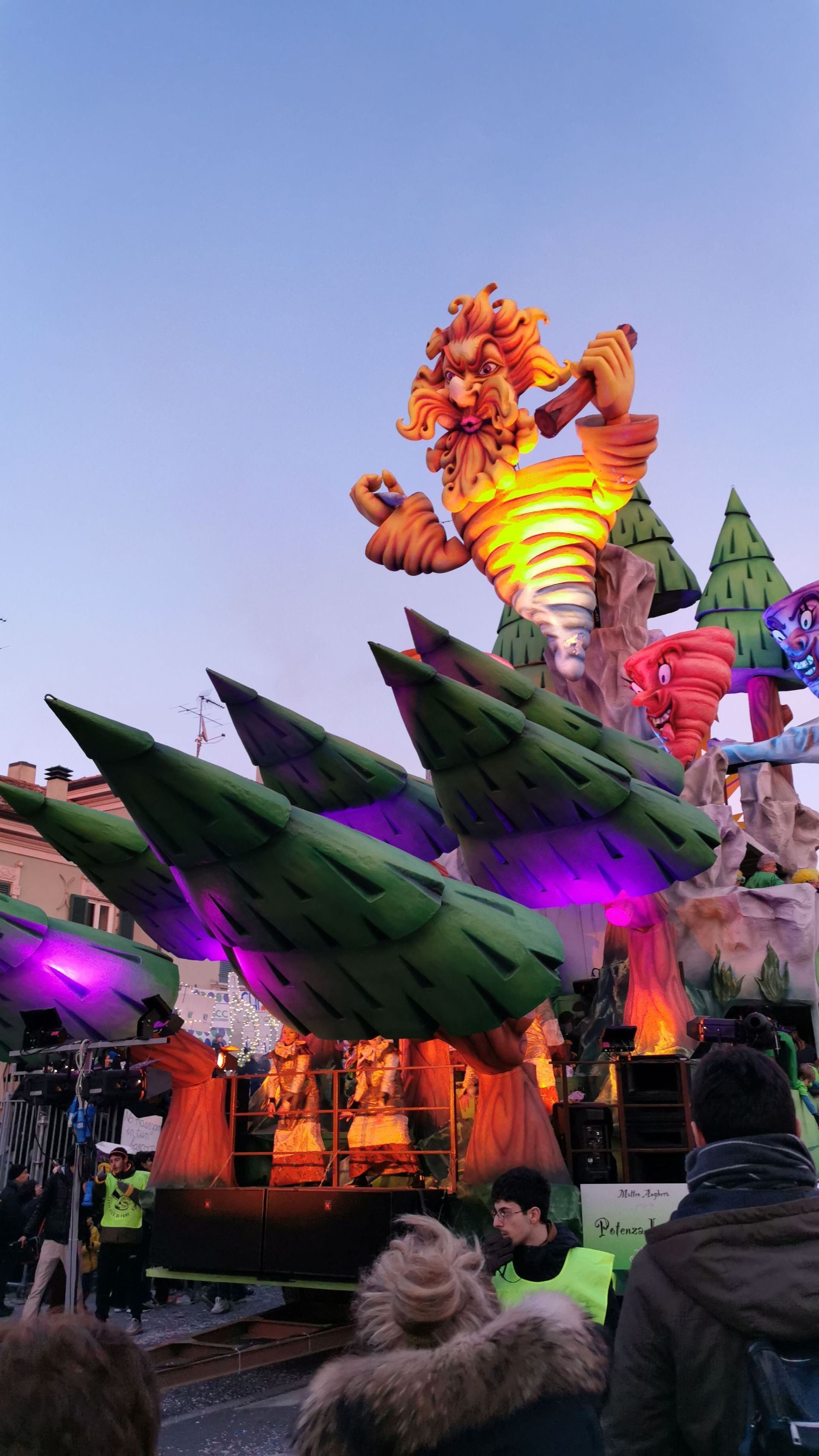 Un grande e colorato carro di carnevale con una gigantesca figura di tornado a cartone animato e alberi stilizzati osservato da una folla al crepuscolo.
