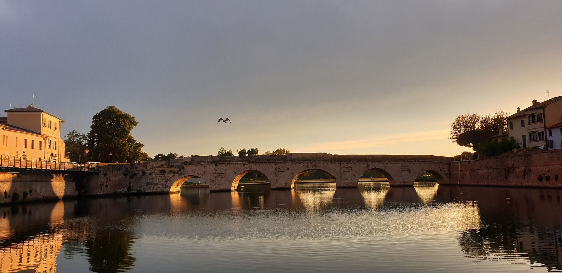 Un ponte in pietra a più arcate che attraversa un fiume calmo al tramonto, con il suo riflesso visibile nell'acqua e un uccello che vola sopra.