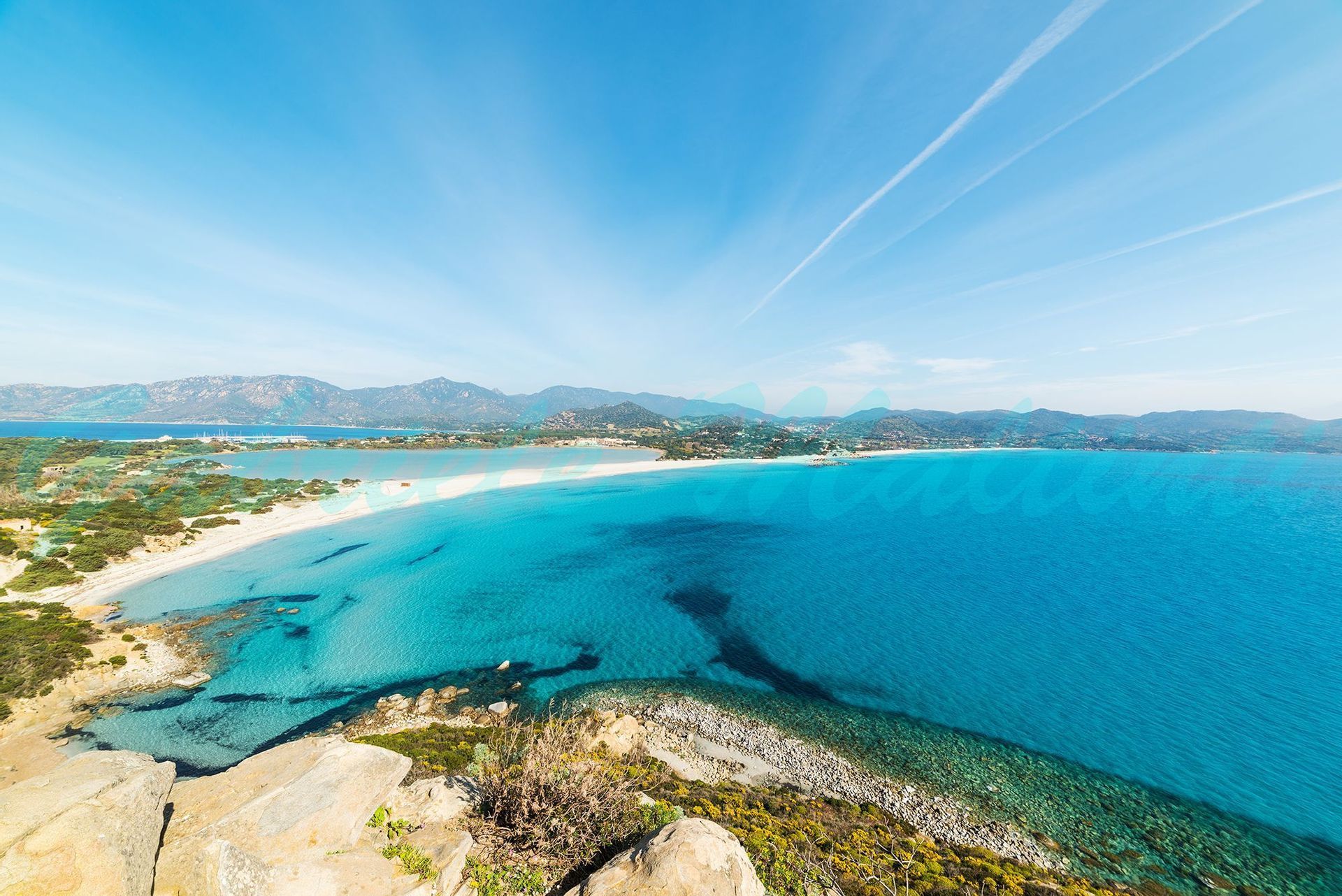 Panoramic view from a clifftop of a white sand beach, clear turquoise water, and distant mountains under a blue sky.