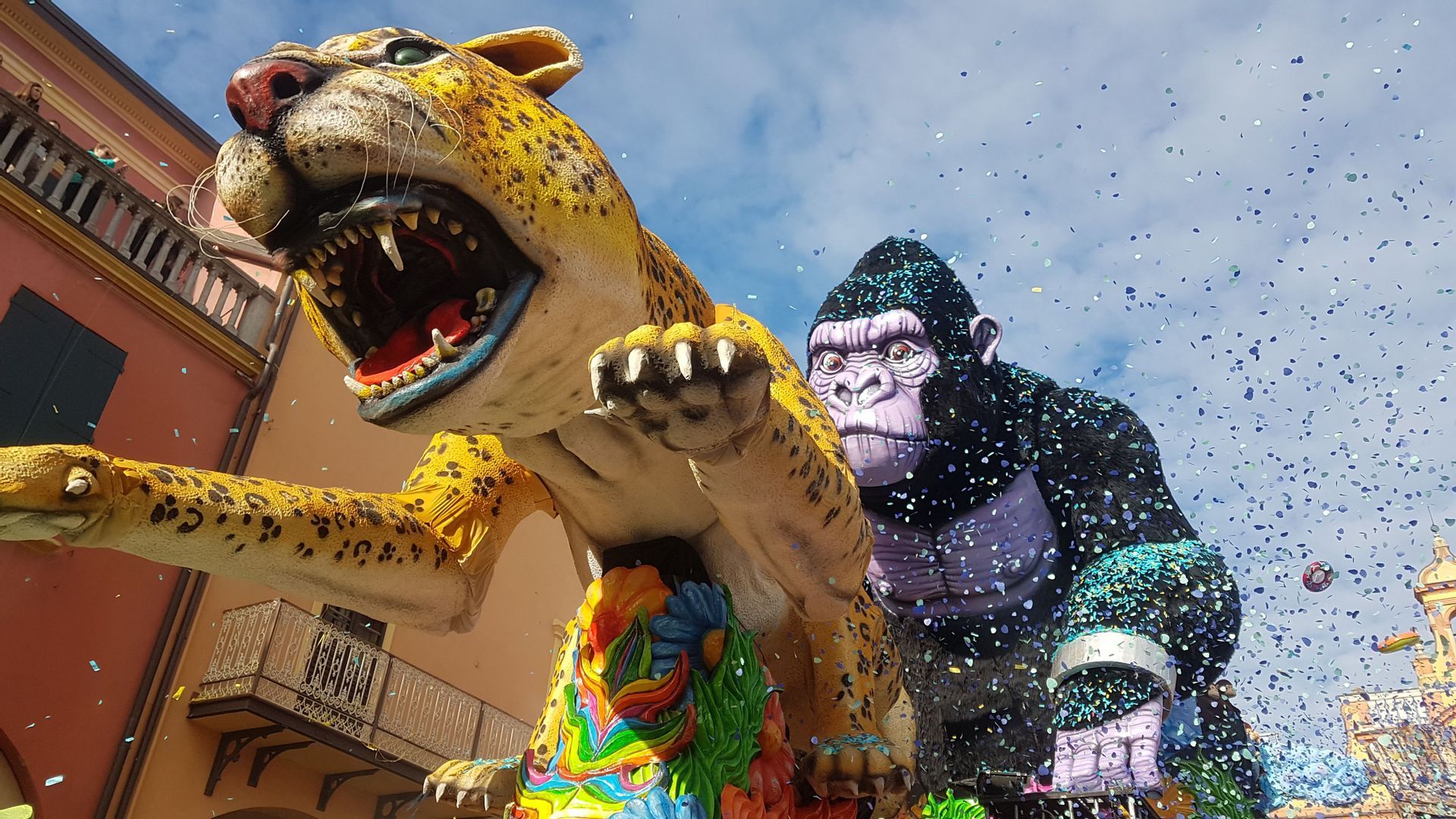 Grandi carri di carnevale con un leopardo ruggente e un gorilla circondati da coriandoli sotto un cielo blu.