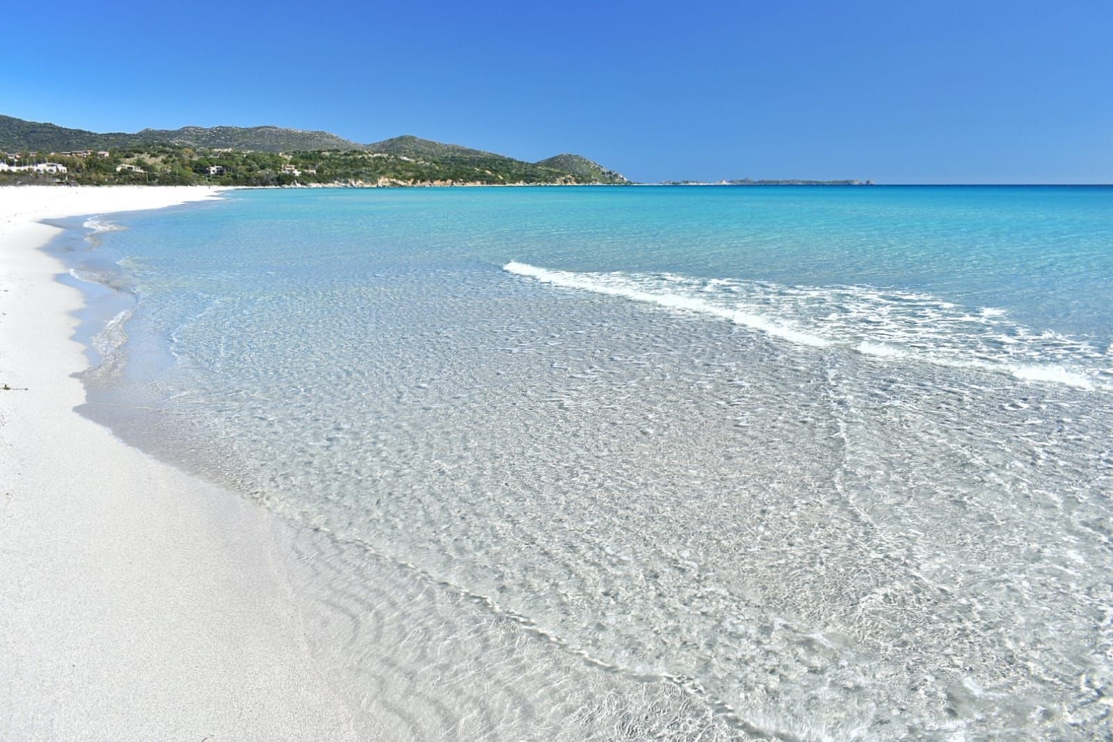 A white sand beach meets clear, turquoise water with gentle waves, set against a backdrop of green hills under a bright blue sky.