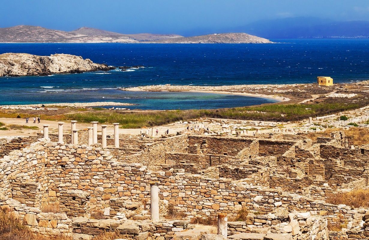 Antiche rovine in pietra con colonne bianche che si affacciano su un mare blu brillante e isole lontane sotto un cielo sereno.