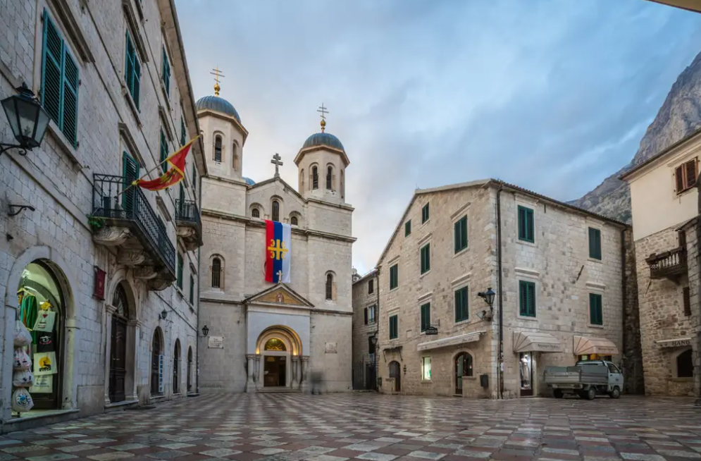 Una chiesa in pietra con doppie cupole si erge in una piazza lastricata a scacchiera, circondata da edifici storici e una montagna.
